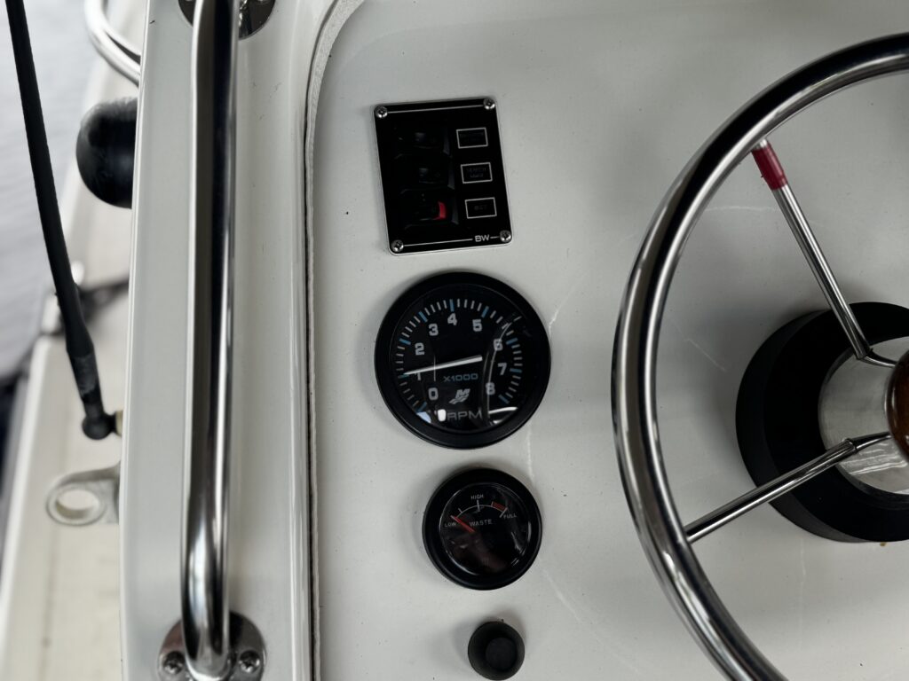 A close-up view of a boat's dashboard, featuring a speedometer on the left, a circular gauge below it, and a control panel with multiple buttons on the upper left. A metal steering wheel with a wooden center is partially visible on the right side.