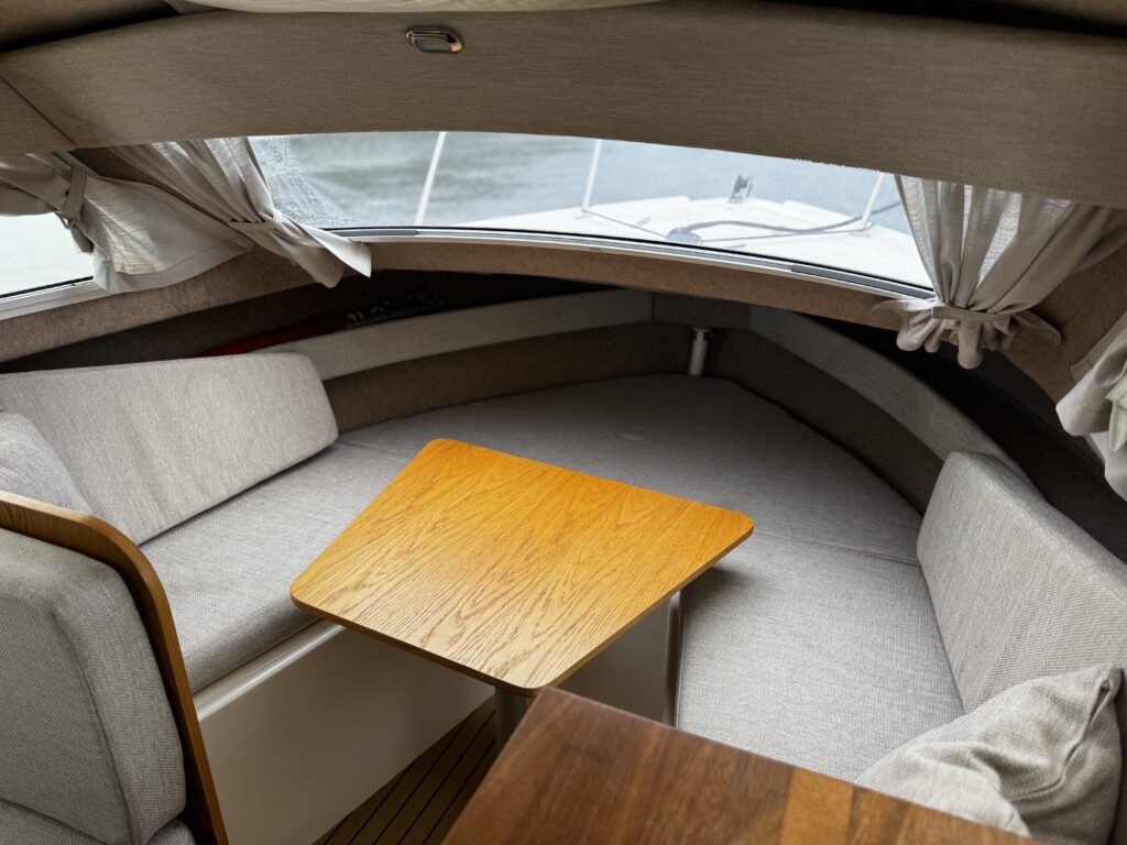 An interior view of a boat featuring a circular seating area with gray upholstered cushions. A wooden table is positioned in the center, complemented by two large windows with white curtains allowing natural light to enter the space.