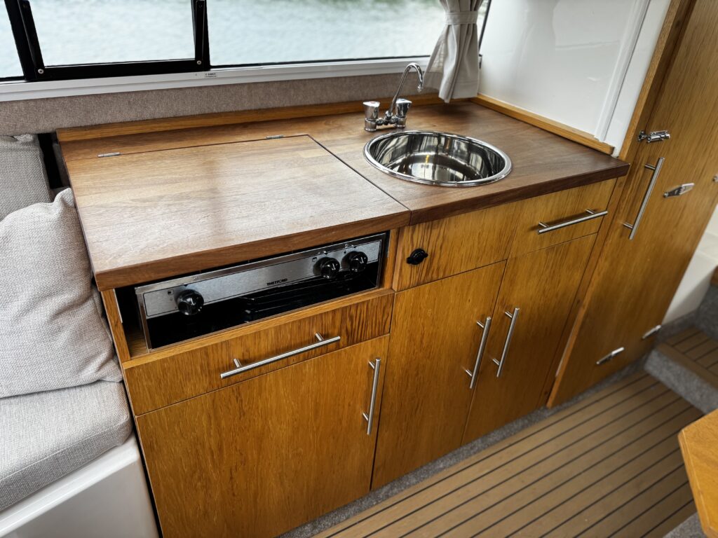 A wooden countertop in a boat galley features a round sink with a faucet and storage cabinets below. An integrated cooking appliance with knobs is visible beneath the countertop, and the interior is well-lit by natural light from a nearby window.