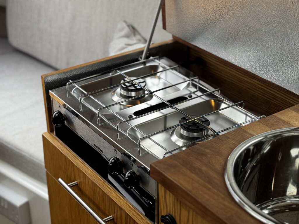 A compact kitchen setup featuring a stainless steel two-burner stove with a protective wire rack above the burners. The stove is installed in a wooden cabinet with a circular sink nearby, and a soft upholstered seat is visible in the background.