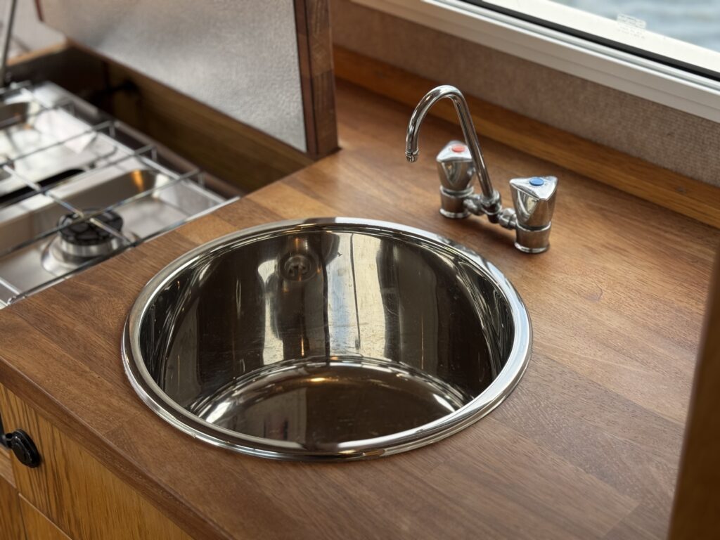 A stainless steel sink with a rounded basin is installed in a wooden countertop. Above the sink is a faucet with hot and cold water controls, and a gas stove is visible on the left side of the image.