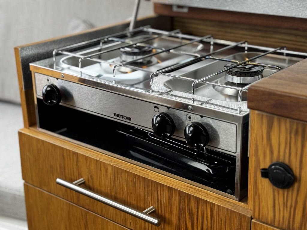 A compact kitchen stove is installed in a wooden cabinet. The stove features two burners with metallic grates and black control knobs, while the cabinet includes a silver handle and a black drawer below.