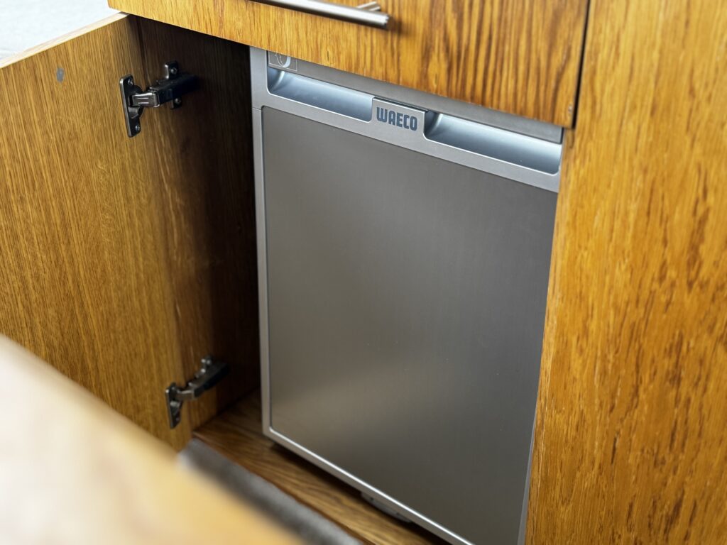 A compact stainless steel refrigerator is built into a wooden cabinet. The cabinet door is open, revealing the refrigerator inside, which features a display panel at the top with the brand name 'Waeco'.