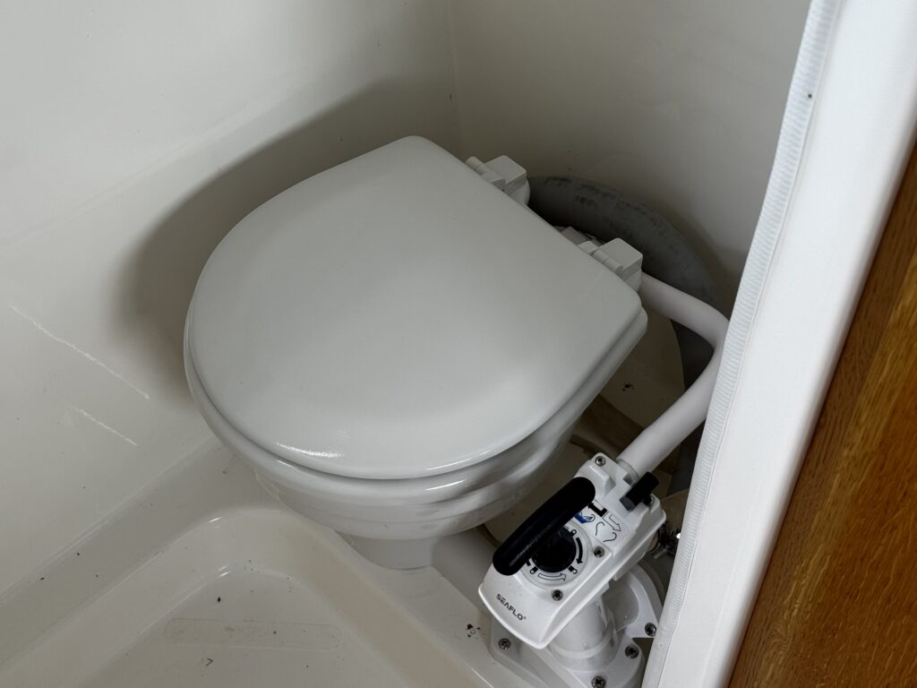 A white toilet situated in a small bathroom corner. The toilet features a control panel on the side, with visible plumbing connections and a plain white interior.