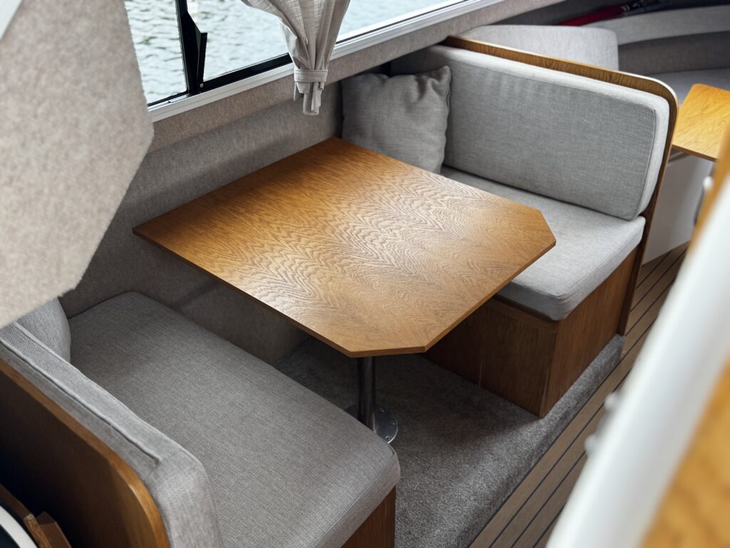 An interior seating area of a boat featuring a hexagonal wooden table surrounded by two cushioned benches. The benches have gray upholstery and are positioned against a window which shows water and reflections outside.