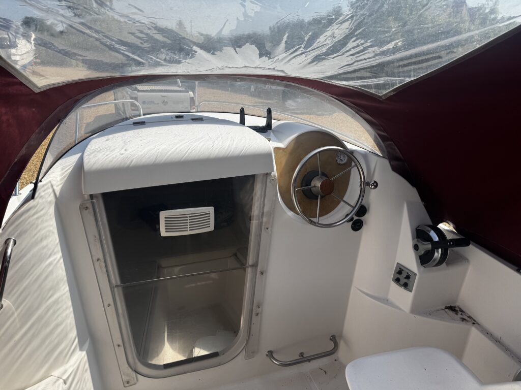The interior of a small boat featuring a steering wheel and controls on the right. A transparent canopy provides visibility, with storage and a white seat visible. The area is predominantly white with maroon accents.