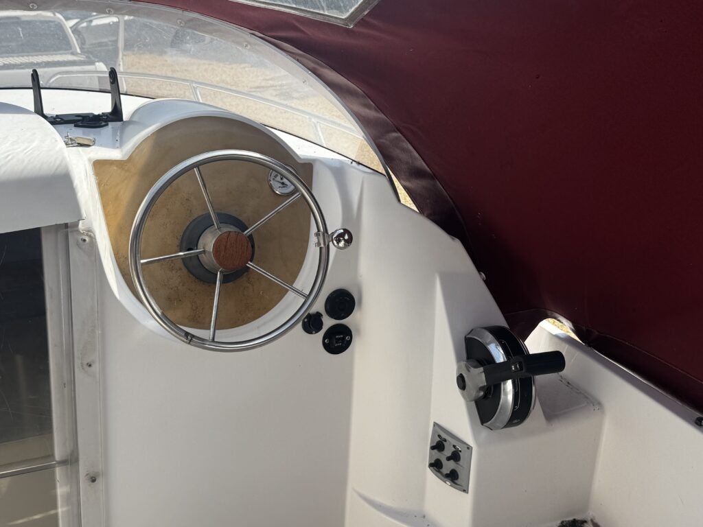 A close-up view of a boat's steering wheel with a wooden center, mounted on a white dashboard. There are various control dials and switches visible, along with a maroon canopy above and part of a window to the left.