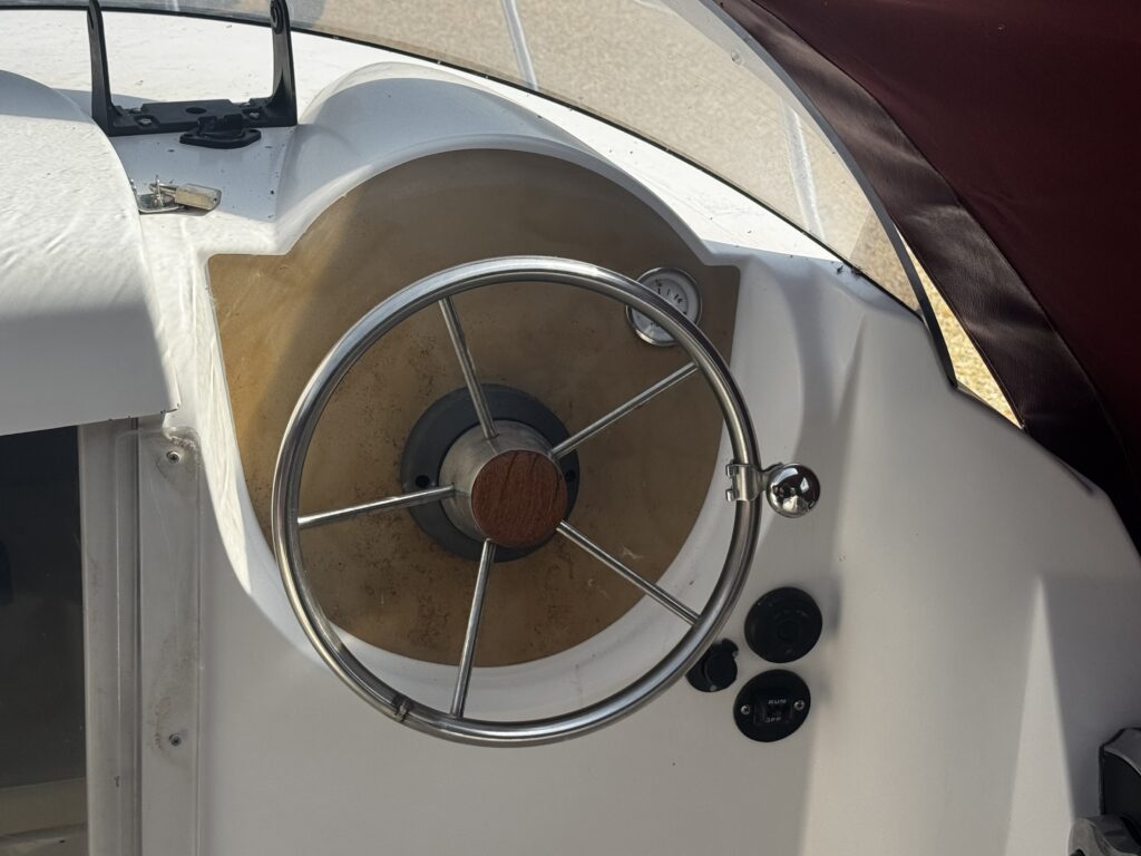 The image shows a boat's steering wheel with a circular chrome frame and a wooden center mounted on a beige panel. Surrounding the wheel are various control buttons, and the background features a white boat interior and maroon fabric.
