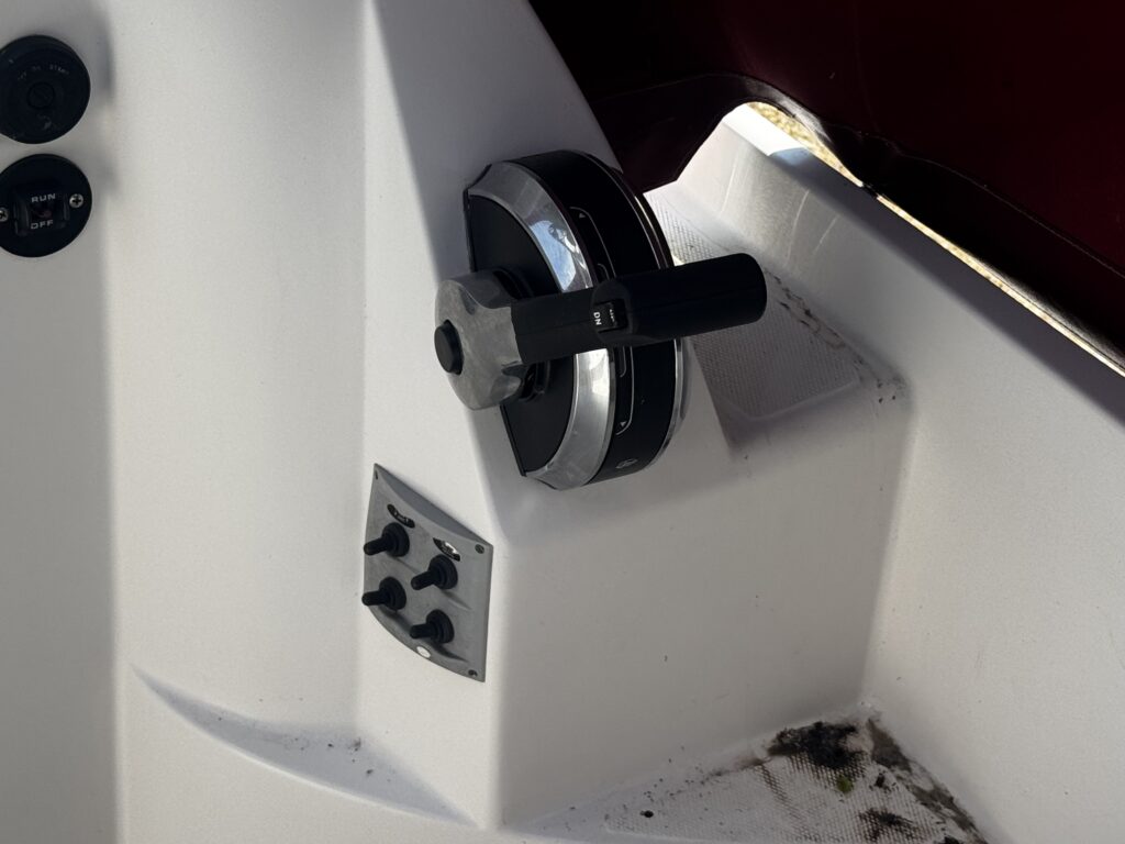 A close-up view of a boat control panel featuring a large black and silver throttle handle. Below the handle are several buttons and switches on a white surface, with hints of dirt in the area.