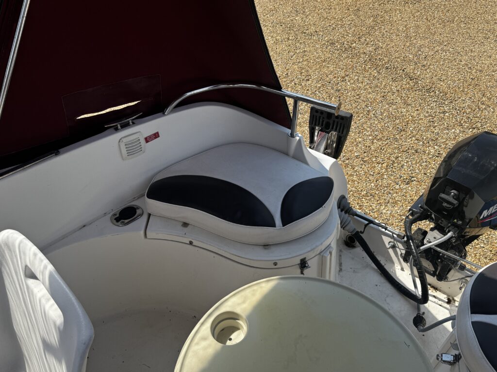 A boat interior showing a white and dark blue cushioned seat beside a control panel. A motor is mounted on the back, and a circular table with a built-in cup holder is also visible. The background has a gravel surface.