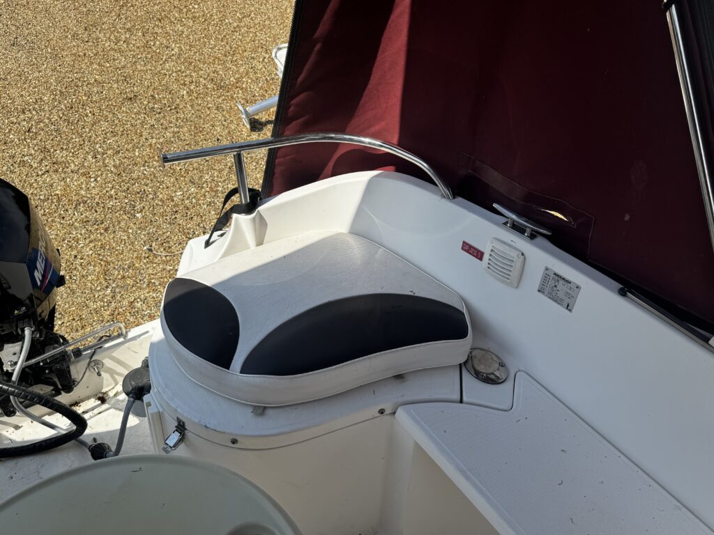 A white and black cushioned seat on a boat, positioned near a control panel. The surroundings include a textured gravel surface and a maroon canopy, with a boat motor visible in the background.