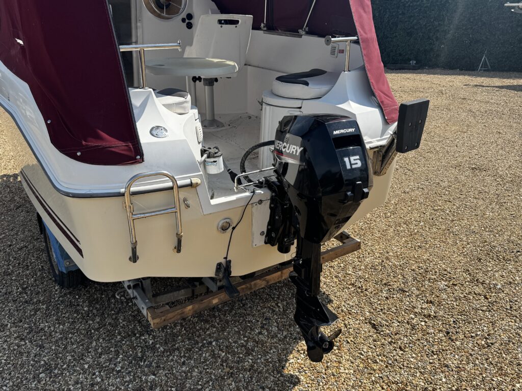 A motorboat with a white hull and maroon canvas cover at the stern, showing a Mercury 15 horsepower outboard motor mounted on a wooden platform. The boat's interior includes seating and an enclosed area.