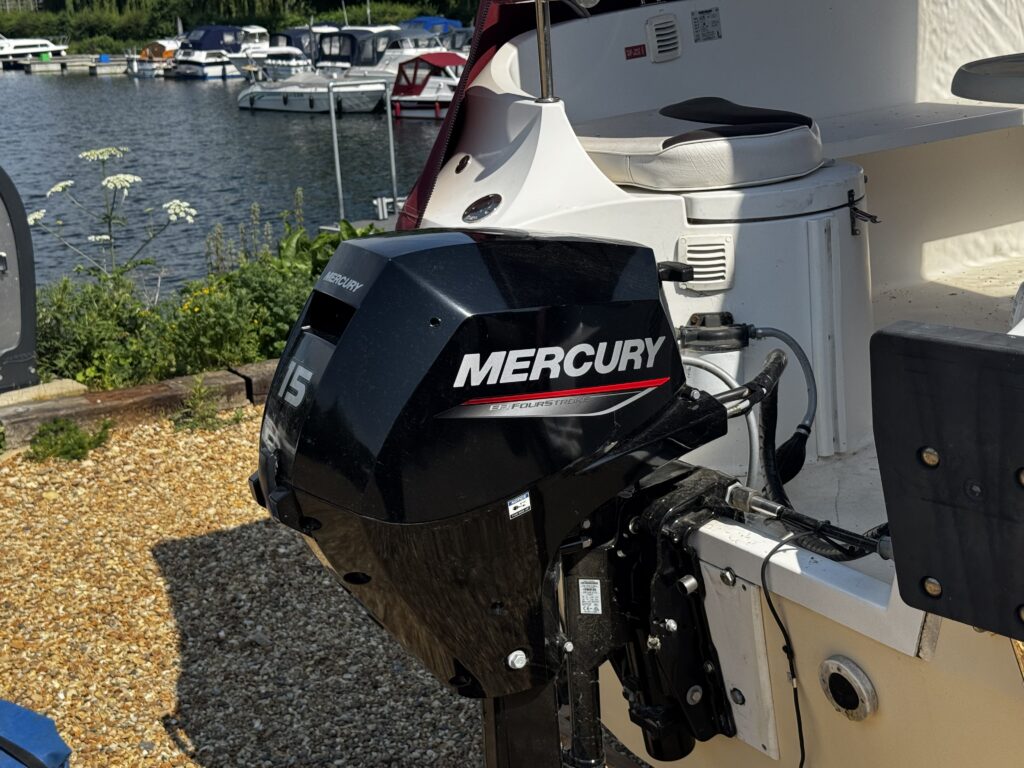 A black Mercury outboard motor, model 15, is mounted on the back of a boat. The background shows a marina with several boats moored on the water, surrounded by greenery and gravel on the dock.