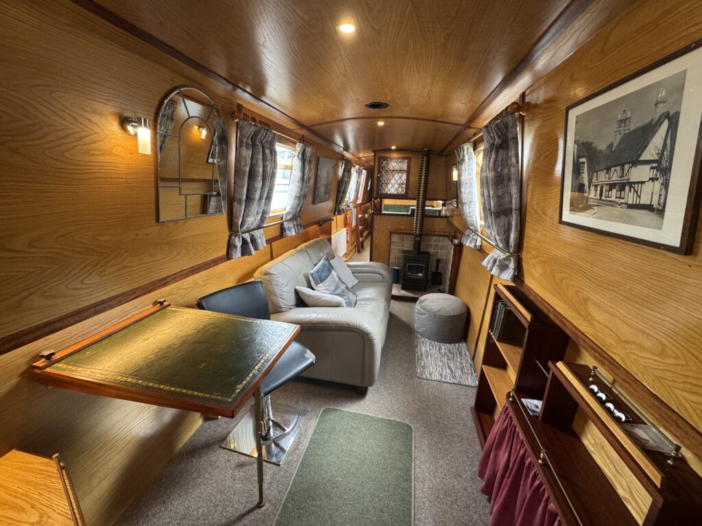 An interior view of a narrowboat featuring wooden paneling, a gray couch with pillows, a small round table, and a plush ottoman. Large windows allow natural light in, and there is a bookshelf and a small stove visible along the walls.