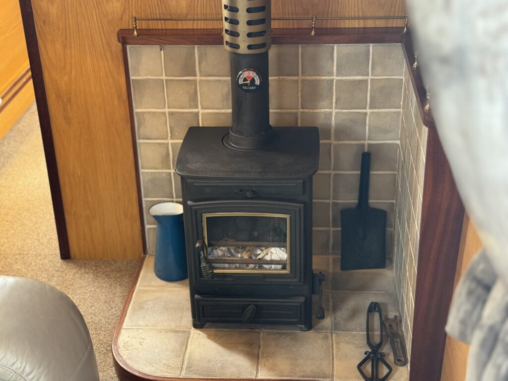 A black wood stove sits against a tiled wall in a room. Next to it, there is a blue pitcher and a black shovel. The area is carpeted and features wooden paneling and a view of a light-colored wall in the background.