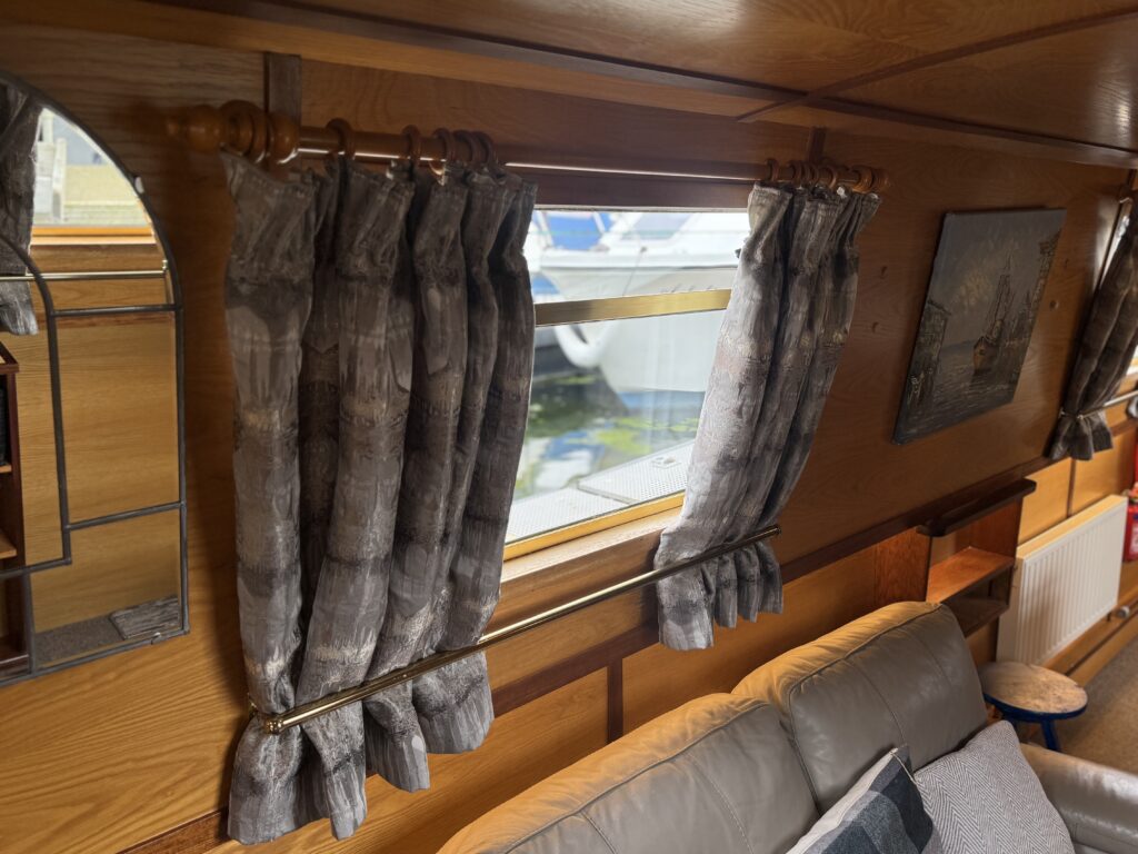 Interior view of a boat featuring gray patterned curtains hanging over a window. The wooden paneling gives a warm feel, with a mirror reflecting light and a painting on the wall. A beige sofa is positioned in front, offering seating.