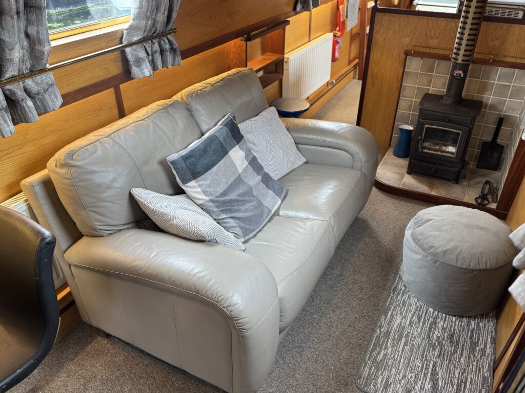 A beige leather loveseat with three decorative pillows is positioned in a cozy interior space. Nearby, a small circular ottoman is on a gray area rug. A wood-burning stove is visible in the background along with wooden paneling and large windows.