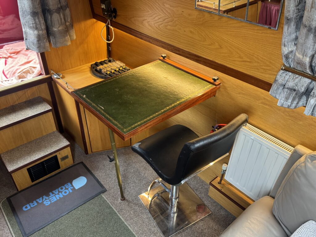 A small workspace in a room featuring a large green desk with brass corners, a black swivel chair, and a radiator next to it. The setup includes wood paneling and a bed in the background, with a doormat displaying a logo.