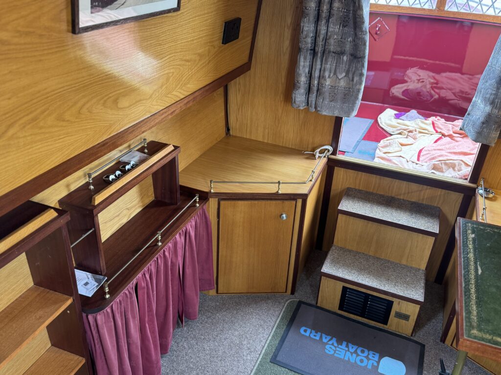 An interior view of a boat cabin featuring wooden paneling, a corner with a small desk, and a cabinet with a red curtain. There are two chairs, a green mat on the floor, and a partially visible window with drapes. A pink blanket is laid on a surface near the window.