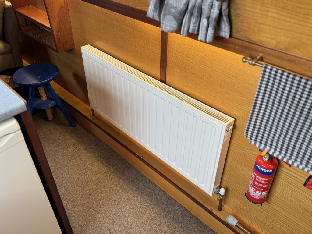 A white radiator mounted on a wooden wall, positioned above a beige carpeted floor. To the left, a blue stool is visible, and a red fire extinguisher is mounted nearby, with a checkered towel hanging on the right side.