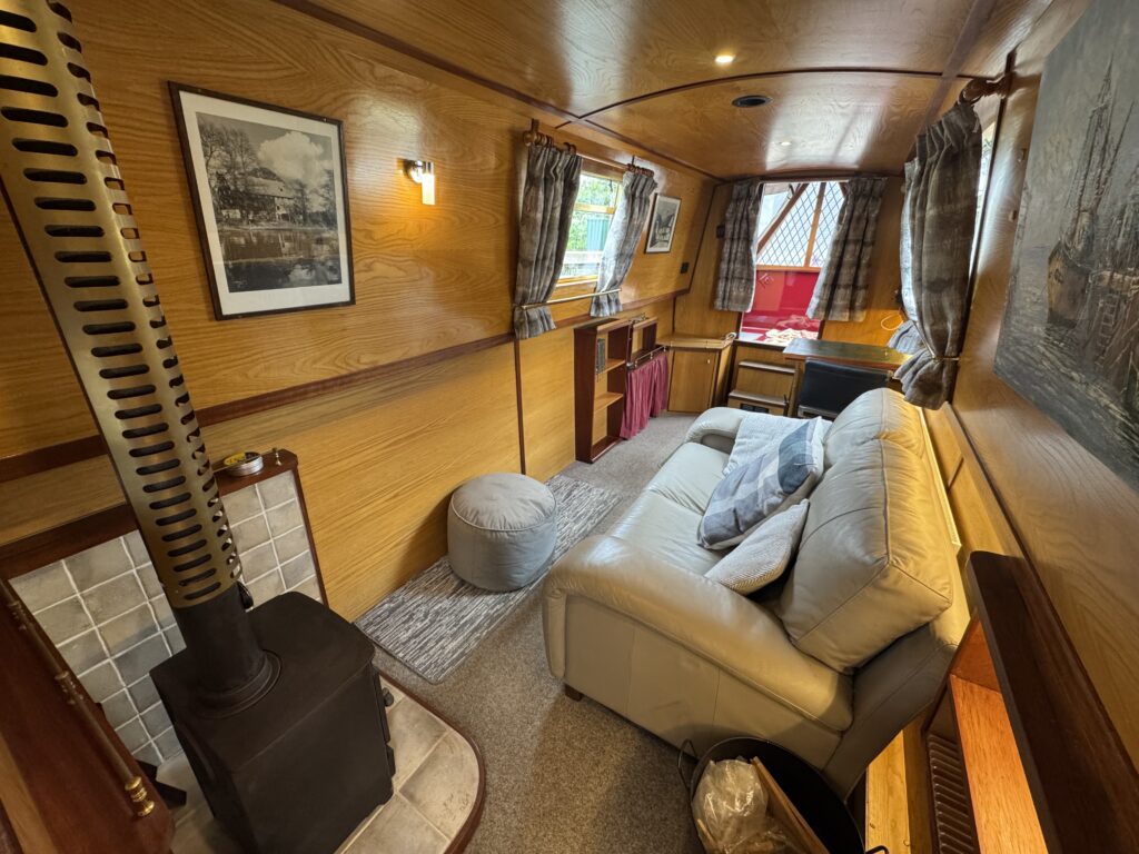 The interior of a narrowboat features wooden paneling, a stove, and a light gray sofa with cushions. There are framed pictures on the walls, large windows with curtains, and a small table beside the sofa, creating a cozy living space.