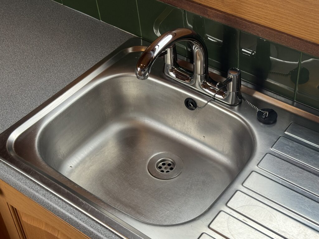 A stainless steel kitchen sink with a sleek, curved faucet and a side rinse sprayer. The sink is set against a green tiled wall and features a visible drain in the center, with a gray countertop and wooden cabinetry surrounding it.
