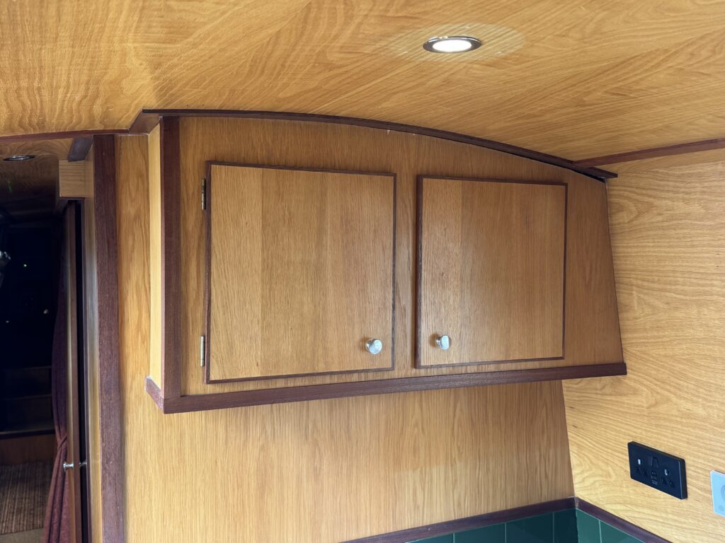 A view of a wooden cabinet with two closed doors and round silver knobs, mounted above a green tiled surface. The ceiling has a recessed light fixture, and the surrounding wooden paneling adds to the warm interior atmosphere.