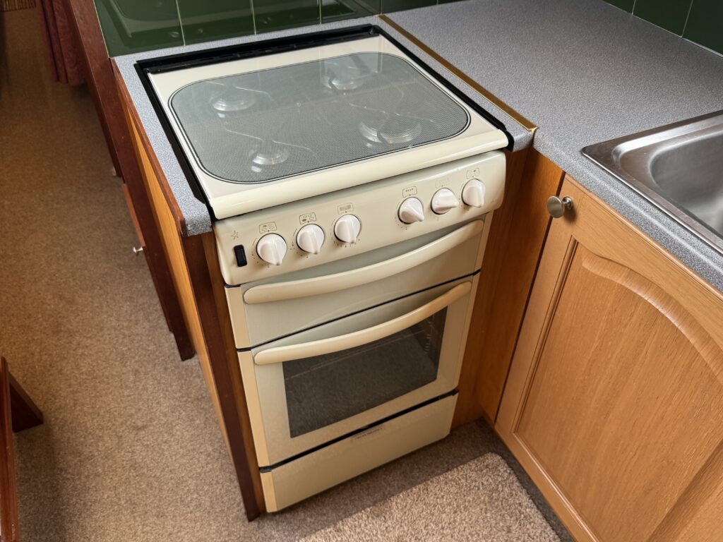 A cream-colored gas stove with four burners on top and an oven below. The stove is located next to a wooden cabinet and a stainless steel sink, set against a backdrop of green tiled walls.