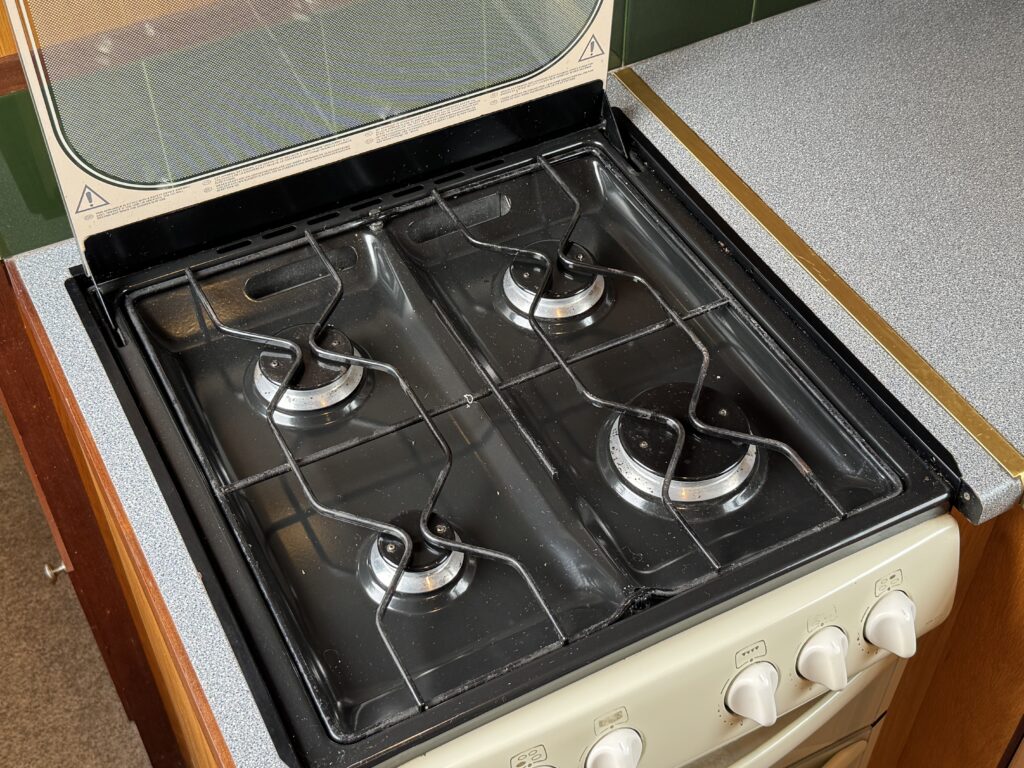 A black gas stove with four burners visible on a countertop. The lid is open, revealing the burners and control knobs located on the front side of the stove. The surface is surrounded by a wooden panel and a gray countertop.