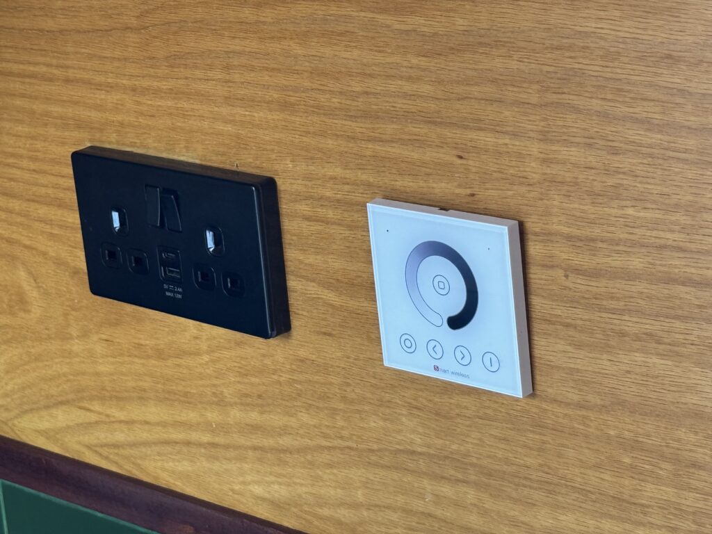 A black electrical outlet panel with multiple sockets and switches is mounted on a wooden wall. Next to it, a white control panel with a circular dimmer dial and four buttons is also attached to the same wall.