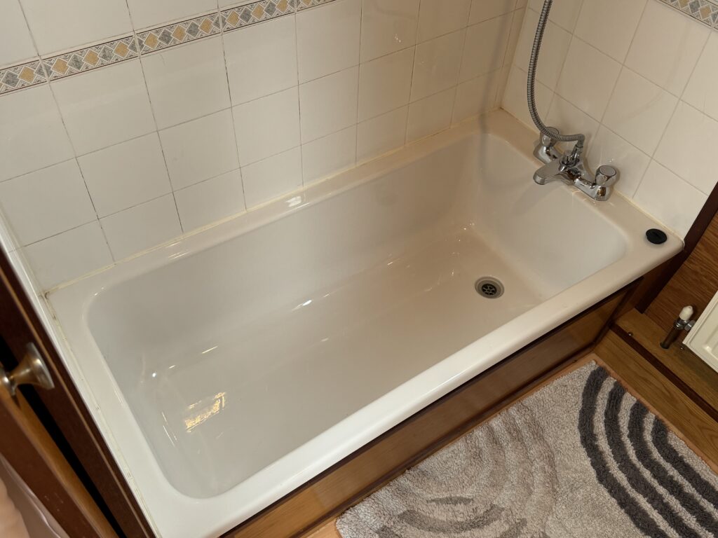 A white bathtub with chrome fixtures and a handheld showerhead. The tiled wall behind features a decorative border, and a plush bath mat with a swirled pattern is visible on the floor.