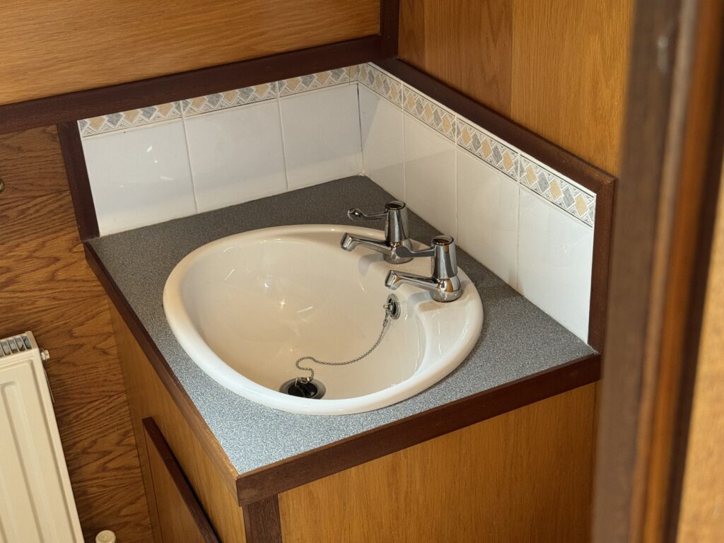 A white circular sink with two silver faucets is installed on a gray countertop. The background features a tiled wall with a decorative border and wood paneling. A small radiator is visible to the left.