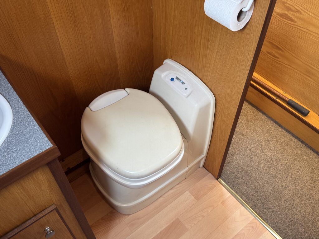A beige toilet with a closed lid, positioned against a wooden wall. A roll of toilet paper is mounted above it. The floor features light-colored laminate, and a corner of a gray sink can be seen to the left.