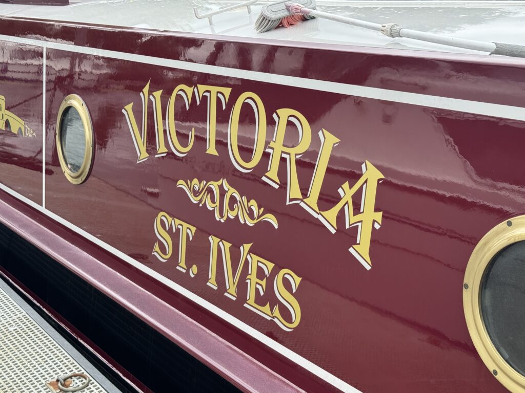 The side of a boat painted in deep burgundy, prominently displaying the name "VICTORIA" in golden, decorative letters, with the words "ST. IVES" below it. The background includes a small circular window and a textured surface of the boat.