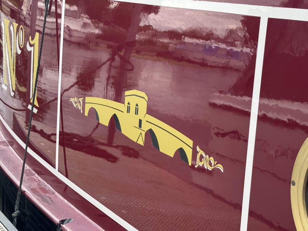 A close-up of the side of a maroon boat featuring a decorative yellow illustration of a bridge, accompanied by ornate detailing. The boat is labeled with the number "1" in a white font above the illustration.