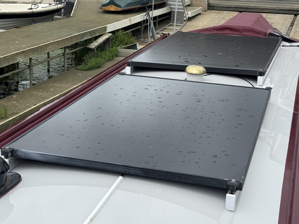 Two solar panels with water droplets on their surface are mounted on a maroon-covered roof. In the background, a marina with boats and a wooden dock is visible, alongside green grass and plants.