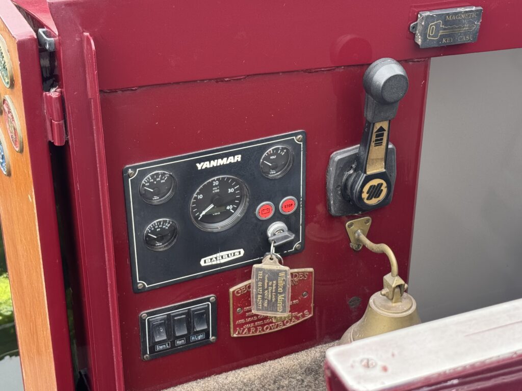 The control panel of a boat featuring a Yanmar gauge with speed and indicator lights, a key ignition, a bronze bell, and various switches. The panel is mounted on a maroon surface with wooden trim, displaying a key and a tag.