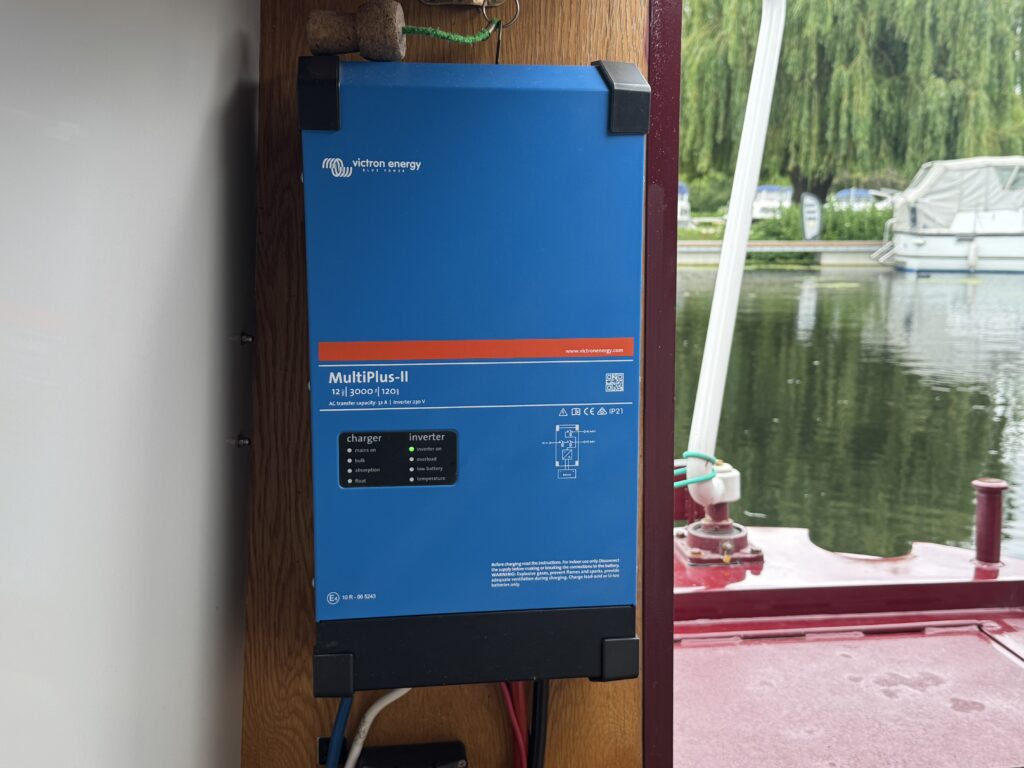 A blue inverter labeled 'MultiPlus-II' mounted on a wooden surface. The inverter has indicator lights and connection ports visible. In the background, boats and trees are partially visible on the water.