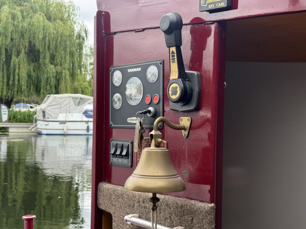 A close-up view of a maroon boat dashboard featuring a Yanmar control panel with displays, control levers, and a brass bell. Reflections of boats and trees are visible on the water in the background.