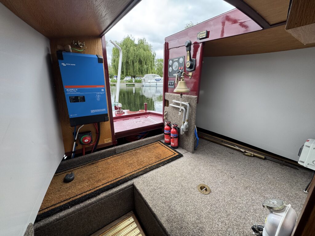 Interior view of a boat showing a blue power inverter mounted on the wall, alongside a gray carpeted floor. Red fire extinguishers are positioned near the water pump, and large windows display greenery and boats in the background.