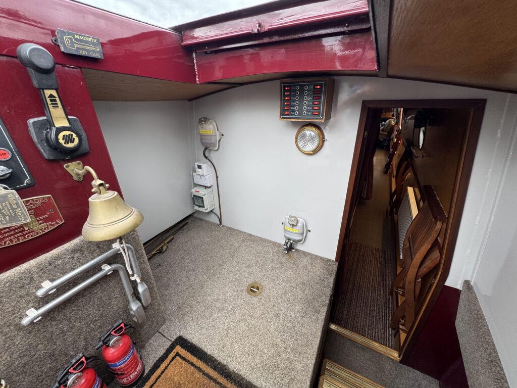 The interior of a boat showing a carpeted area with a bell, fire extinguishers, and several control panels on the wall. A wooden door leads to another area of the boat, and the space is well-lit with a small window.