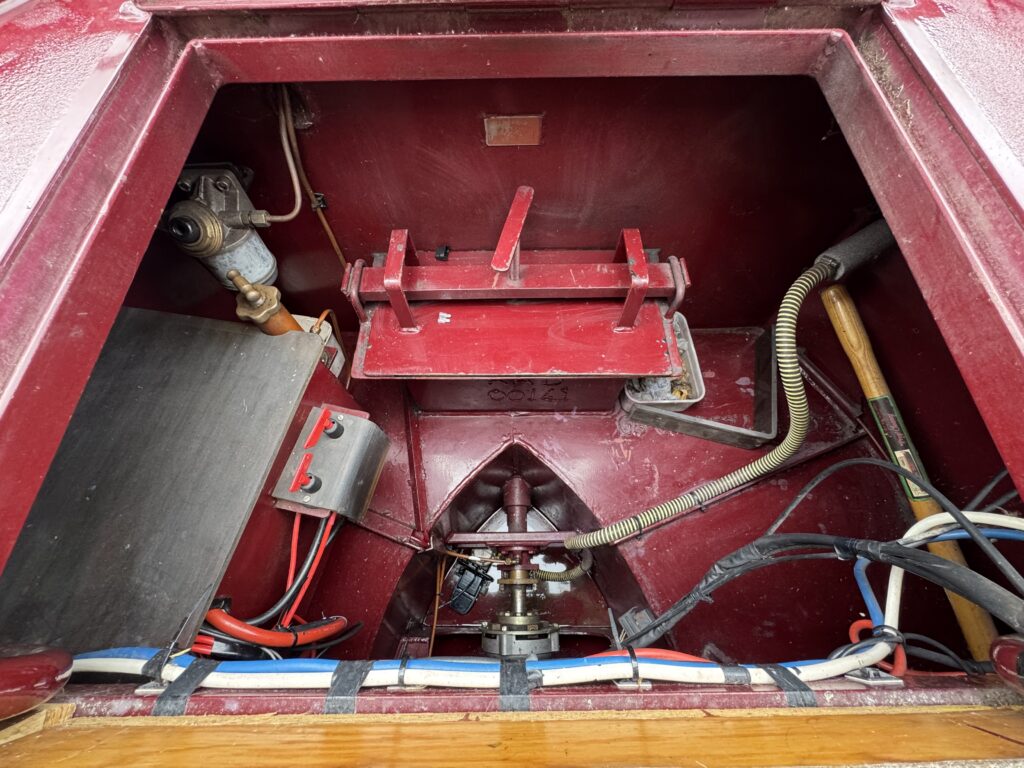 An interior view of a boat's engine compartment showing various mechanical components, including a red metal mounting frame, wires, a battery, and pipes. The compartment has a red finish with wooden edges surrounding the opening.