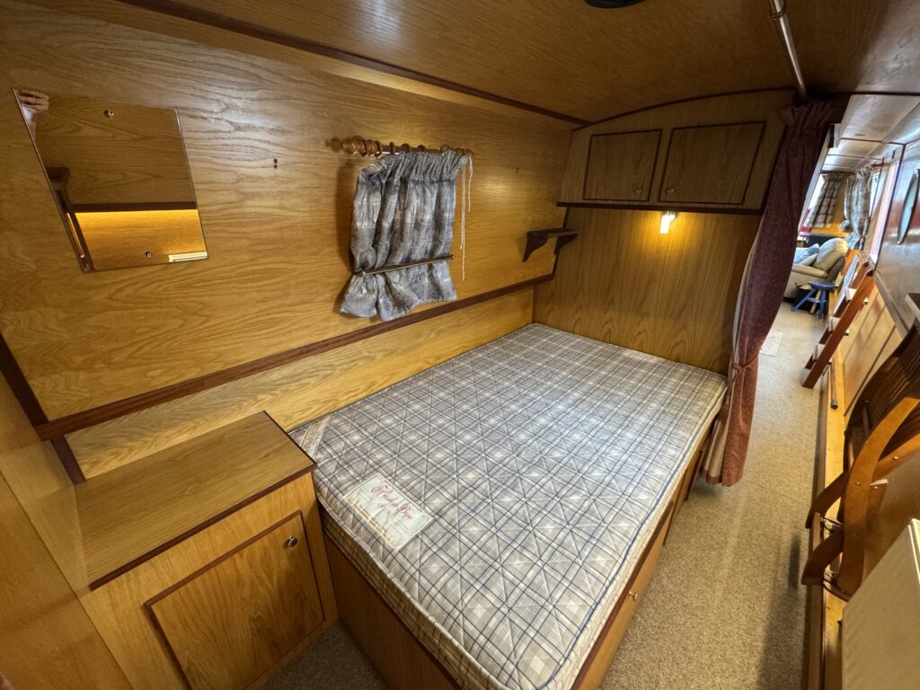A compact bedroom with wooden paneling features a double bed with a plaid duvet cover, a bedside table with an open compartment, and a small mirror mounted on the wall. A window with a patterned curtain provides light, and the room has storage cabinets.