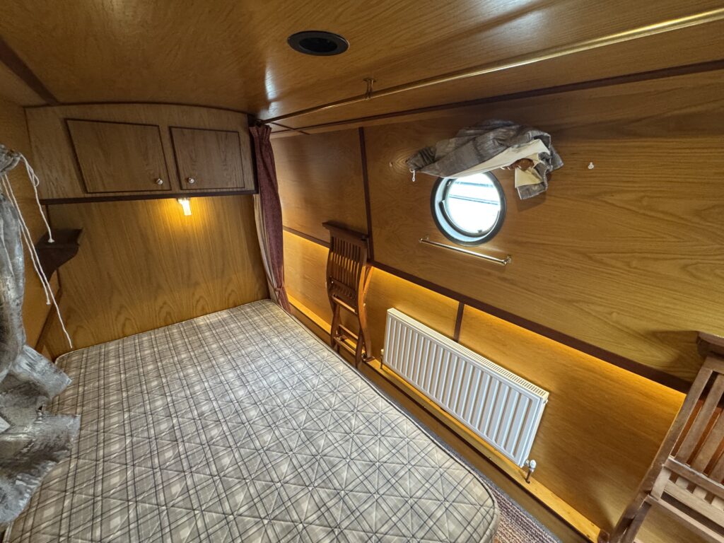 An interior view of a cabin with wooden walls and a double bed covered with a plaid blanket. A small round window is visible on the wall, along with a radiator beneath it, and a wooden chair placed beside the bed.