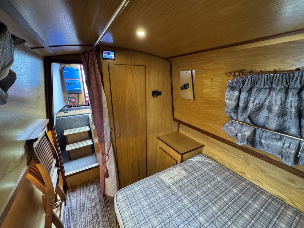 An interior view of a compact room featuring a bed with a plaid mattress and wooden frame. To the left, there is a ladder leading to a door, and the walls are lined with wood paneling. A window with curtains is visible, letting in light.
