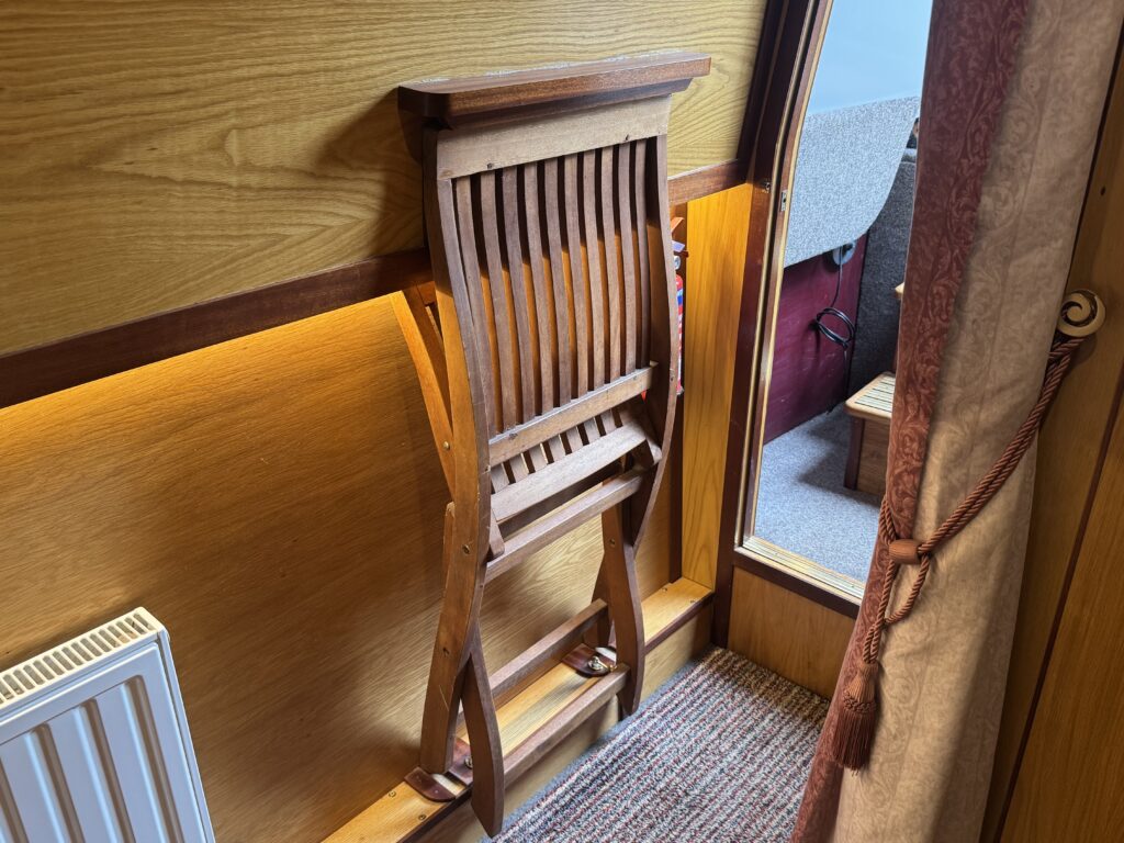 A wooden folding chair stacked against a wooden wall. The chair has slatted backing and a light finish. A portion of the floor shows carpeting, and a radiator is visible along with a partially visible room beyond the doorway.