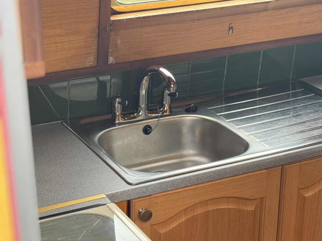 A stainless steel sink with a single tap is installed in a kitchen setting. The countertop is gray with a silver dish drying rack beside the sink, and green tiled backsplash above the counter. Wooden cabinetry is visible in the background.