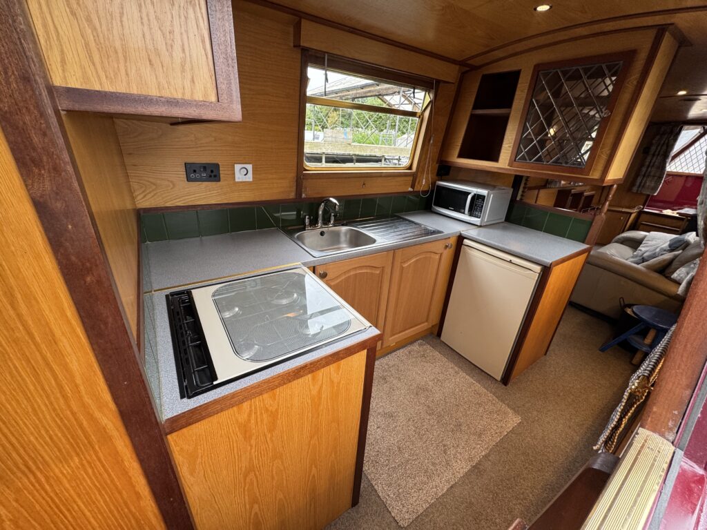 A small galley kitchen featuring wooden cabinetry and a countertop with a sink. On the left, there is a stove, and a microwave is placed above a refrigerator on the right. Natural light enters through a window, with a small rug on the floor.
