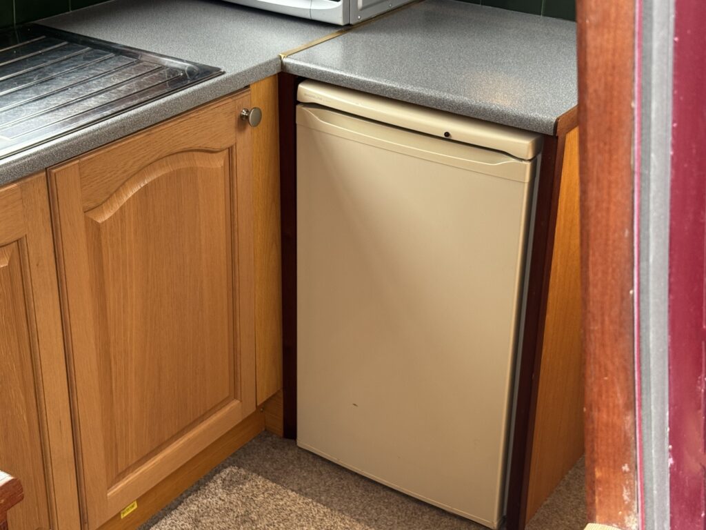 A small beige refrigerator is installed under a countertop, next to a wooden cabinet with raised panel doors. The kitchen has a sink with a metal draining board on the left side, and the countertop is gray. Natural light illuminates the scene.