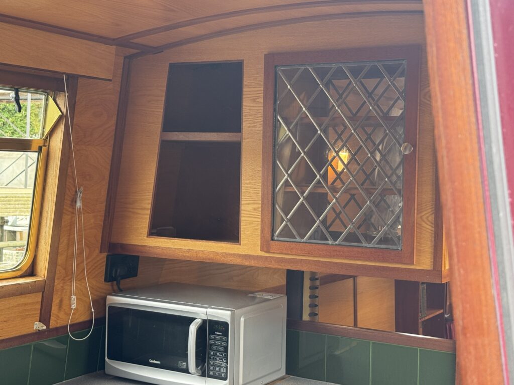 An interior view of a narrowboat kitchen area featuring a microwave on a green tiled surface. Above the microwave, there is a wooden cabinet with glass doors, and to the left, a window allows natural light to enter the space, reflecting off the wooden paneling.
